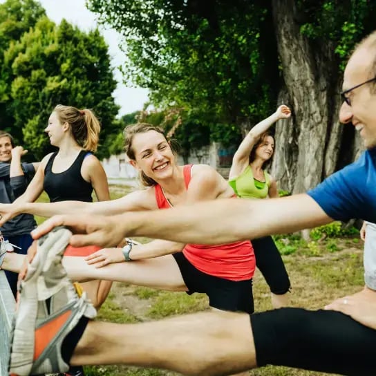 A community of people smiling and stretching their legs on a fence outdoors, surrounded by green trees and grass on a sunny day, enjoying movement that supports their mental health.