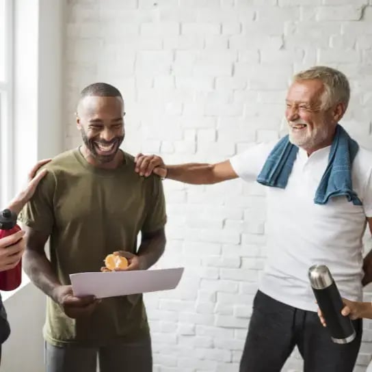 Two people in athletic wear smile and chat indoors, highlighting employee wellbeing benefits. One holds a water bottle as the other carries a sheet of paper and a peeled orange. A third hand rests on the mans shoulder, suggesting friendly interaction.