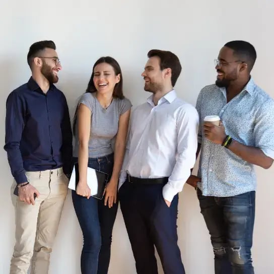 Four young adults stand against a white wall, smiling and laughing together. Their positive energy reflects a happier & more productive workforce—one man holds a coffee cup, another wears glasses, while a woman with a notebook adds to the fitter team vibe.
