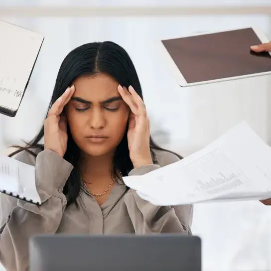 A woman sits at a desk with her eyes closed and hands on her temples, showing signs of hr burnout as multiple hands offer her documents and a tablet, highlighting overwhelming work or information.