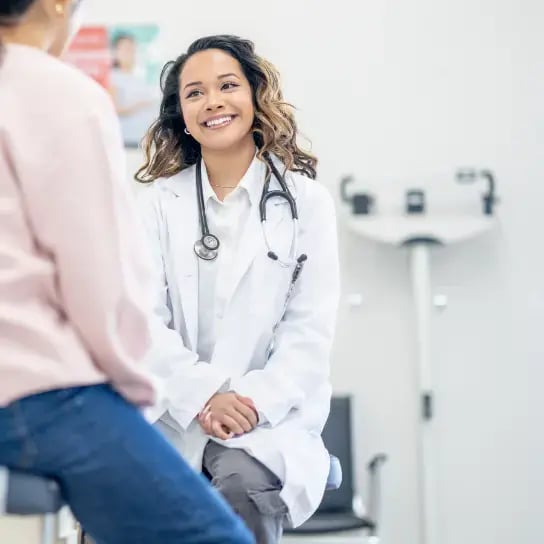 A doctor in a white coat with a stethoscope sits across from a patient in a medical office, engaged in conversation during a health assessment.