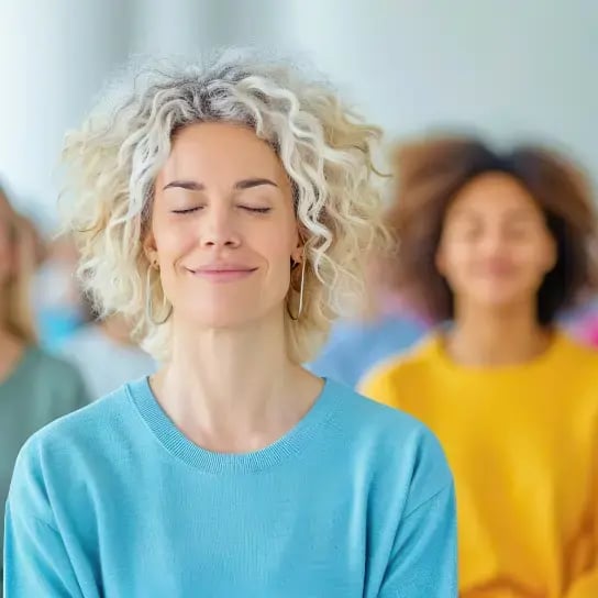 A woman with curly blonde hair and a blue sweater smiles with her eyes closed, appearing relaxed—an example of supporting employee mental health with exercise. Several people are blurred in the background, also sitting calmly with eyes closed.
