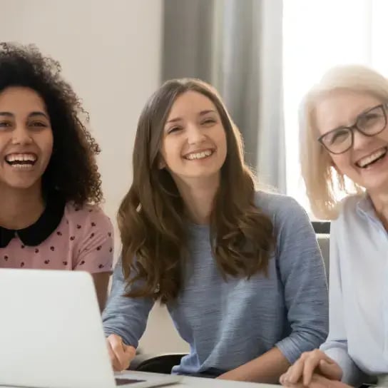 Three women of different ages sit together indoors, smiling and laughing with a laptop in front of them. They appear happy and engaged, reflecting a collaborative atmosphere that highlights supporting women in the workplace.
