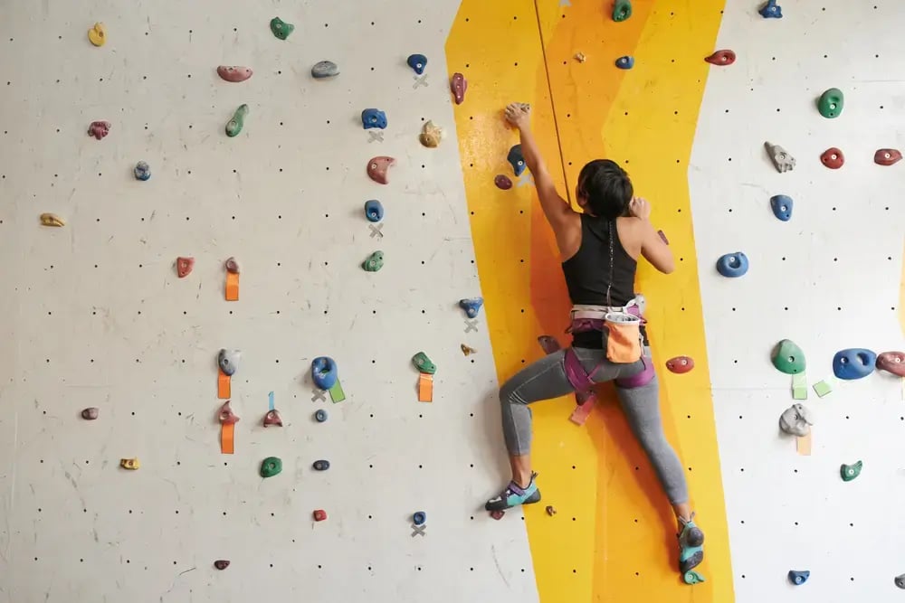 A person wearing a harness climbs an indoor rock climbing wall with colourful holds, focusing on a bright yellow route—an engaging activity that promotes employee wellbeing through physical and mental challenge.