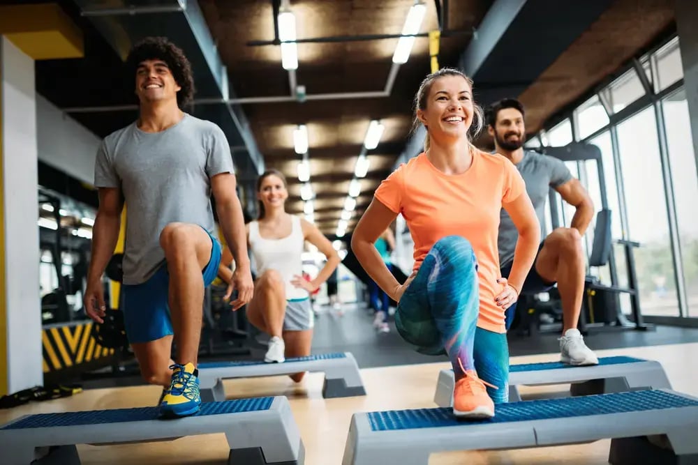 A group of four people exercise in a gym, smiling while performing step-up lunges on aerobic steps. Bright lights and gym equipment are visible in the background, highlighting the great atmosphere and discounted gym memberships available.