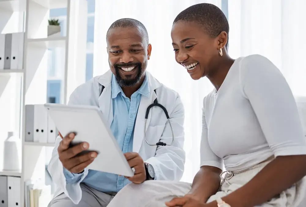A smiling doctor shows a tablet to a cheerful woman during a consultation in a bright, modern office, highlighting their focus on employee wellbeing. Both appear engaged and comfortable.