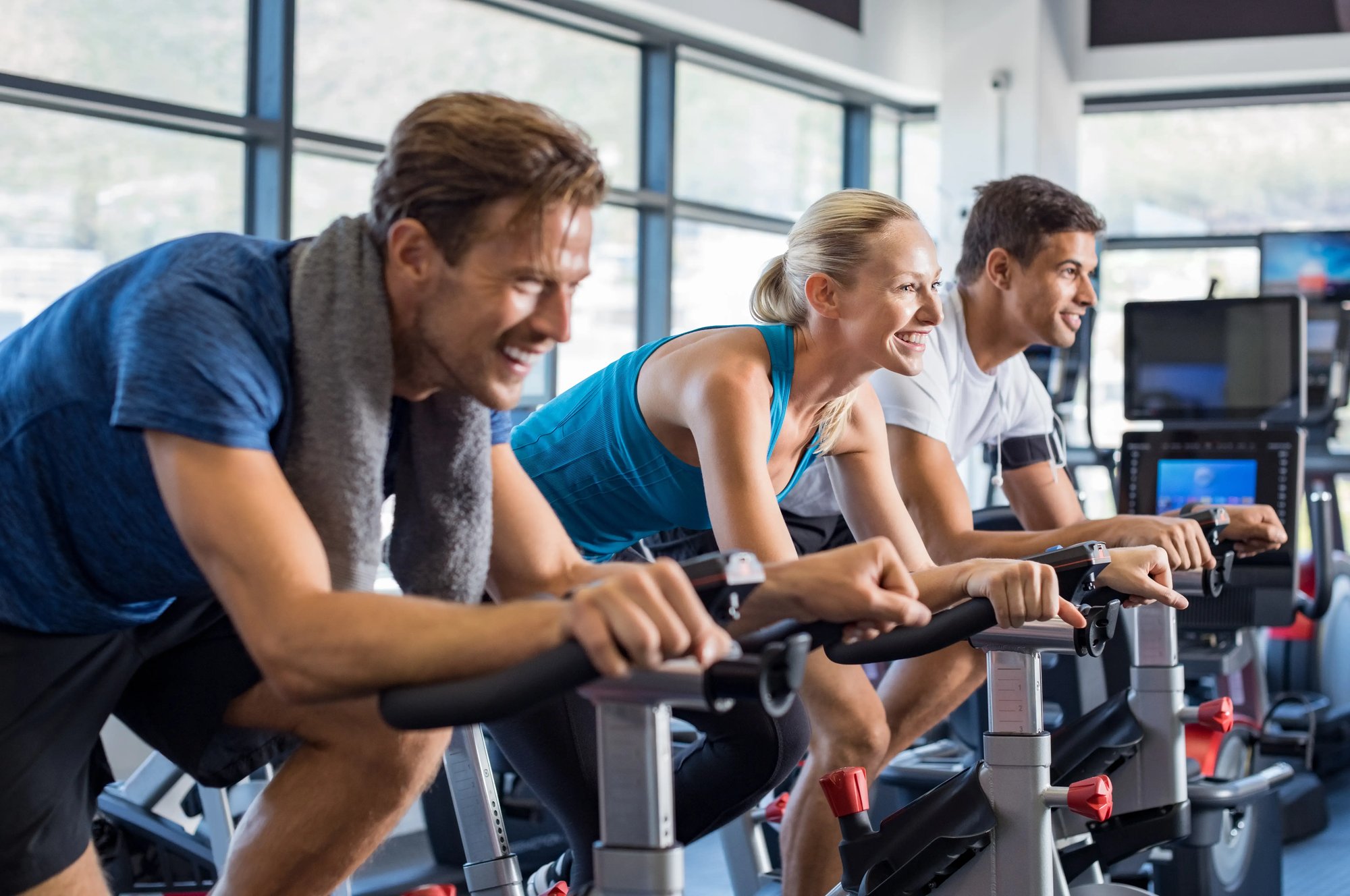 Three smiling adults, two men and one woman, ride stationary bikes in a bright gym with large windows. They appear energetic and focused, enjoying a group indoor cycling workout as part of their corporate gym memberships.