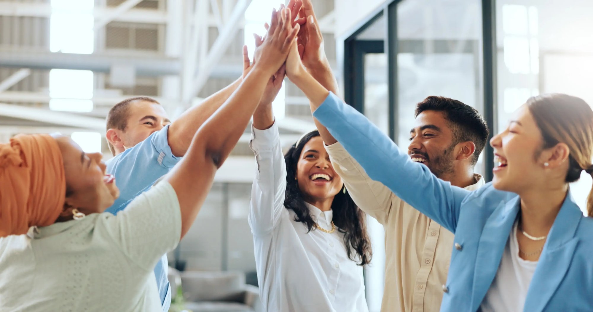 A diverse group of five people stand in a circle and smile as they give each other a high five, celebrating together in a modern office after participating in corporate health assessments.