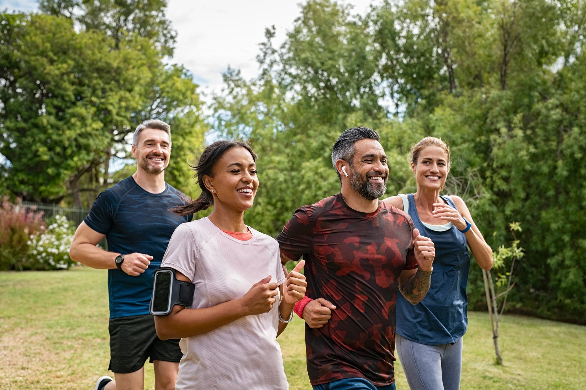 A group of people running cheerfully in a park, surrounded by trees and grass under a bright sky. The scene captures the joy of an active lifestyle with diverse individuals including men and women.