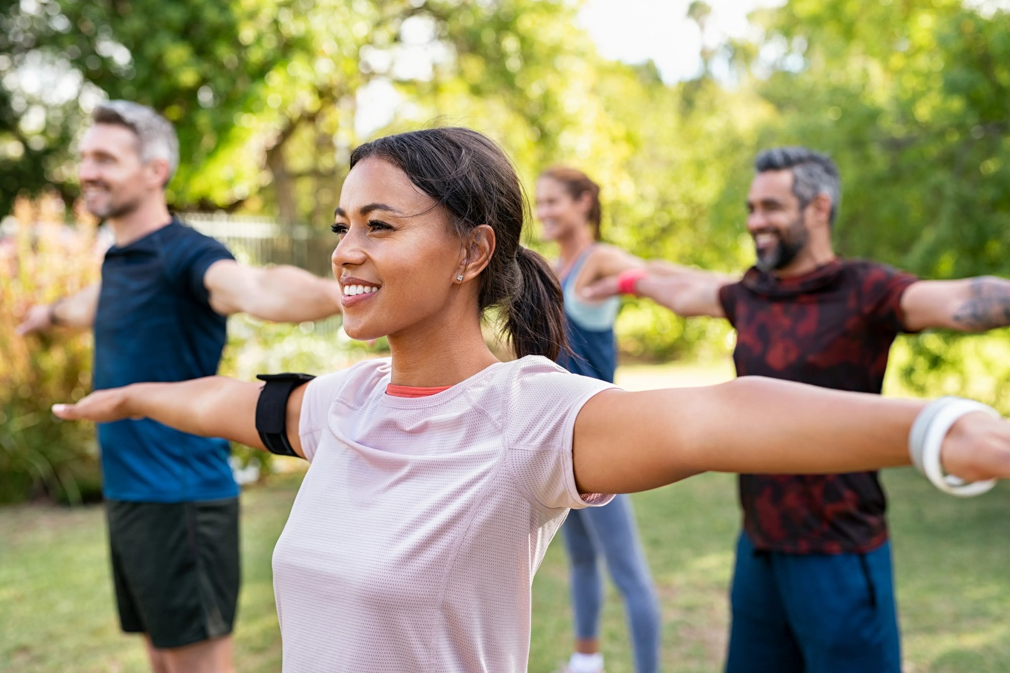 A group of people stretching in a park, surrounded by trees and grass, wearing sports clothing and smiling.