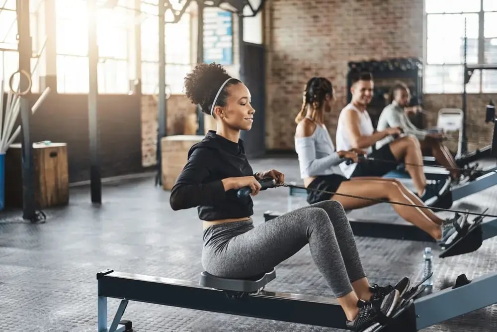 Four people are exercising on rowing machines in a gym with large windows and exposed brick walls, highlighting a workplace dedicated to employee wellbeing. The focus is on a woman in the foreground wearing a black hoodie and gray leggings.