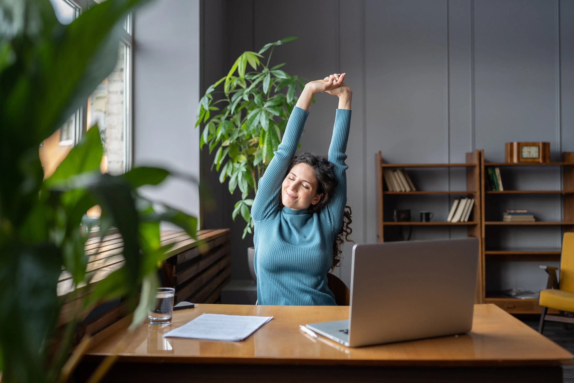 A woman taking care of her health is stretching her arms in front of a laptop on a desk, surrounded by indoor furniture, a book, and a houseplant.
