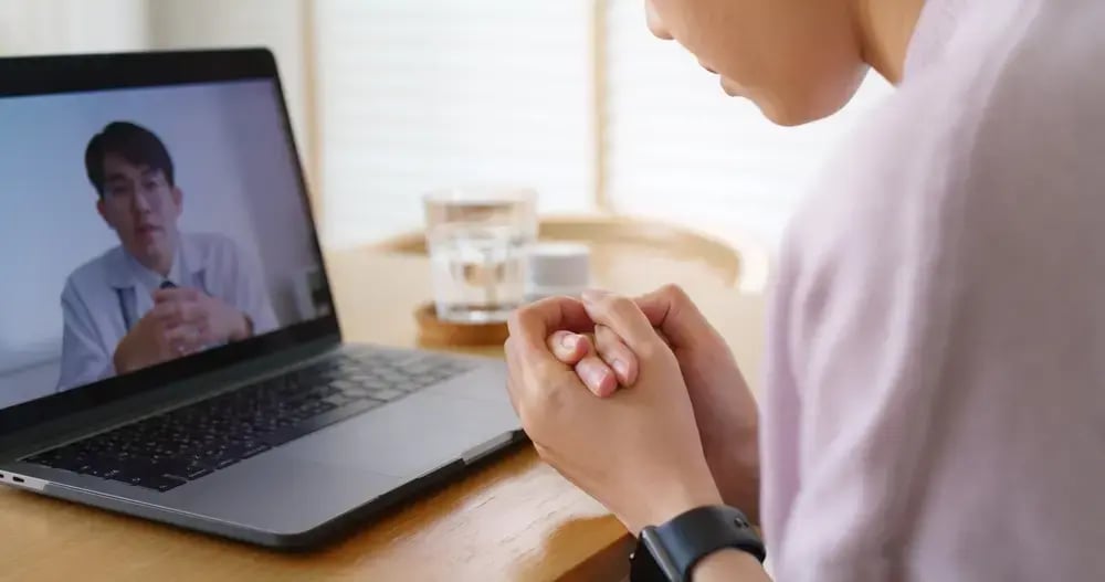 A person sits at a table, clasping their hands, engaged in an online counselling session with another person on a laptop screen. A glass of water and some items are on the table next to the laptop.