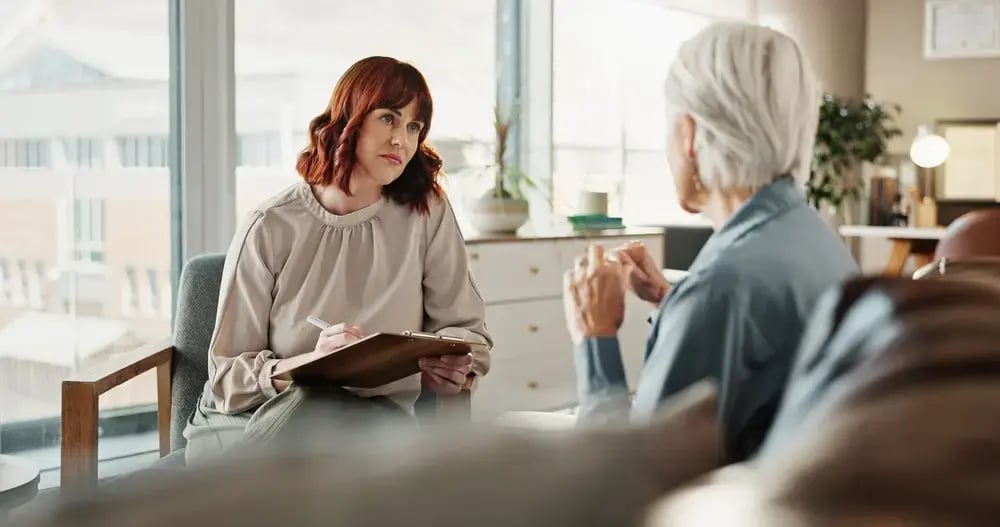 A woman with red hair listens attentively and takes notes on a clipboard during an in-clinic health assessment while sitting across from an older person in a bright, modern office setting.