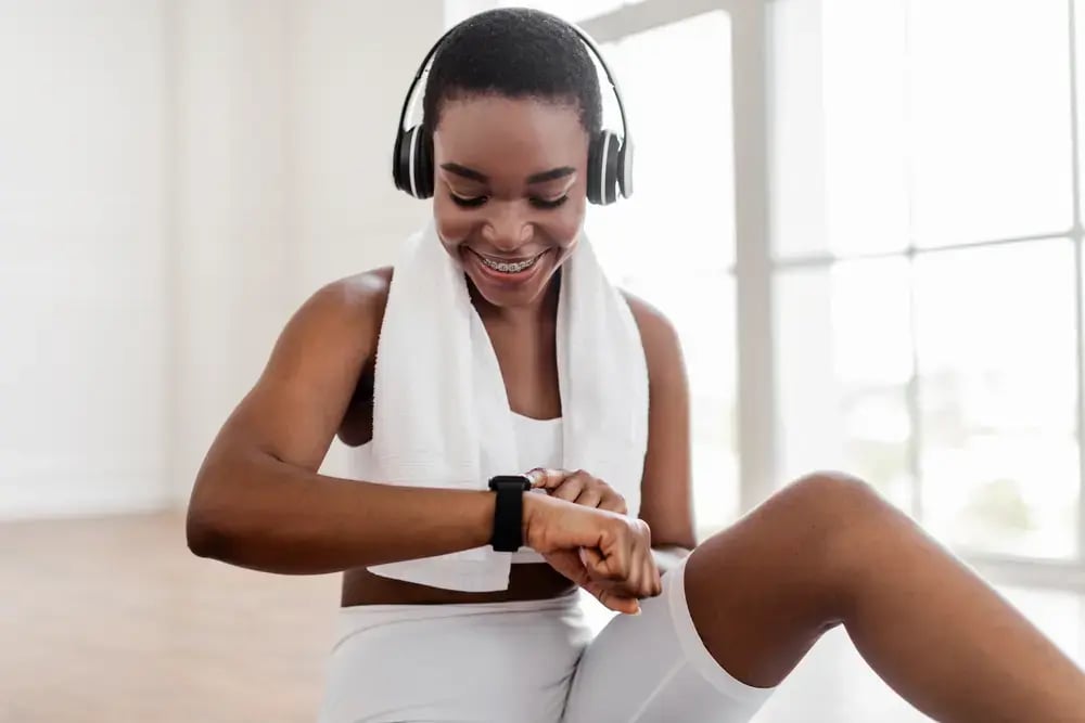 A smiling woman wearing headphones and workout clothes sits indoors with a towel around her neck, looking at her smartwatch - reflecting on her fitness progress after a productive workout.