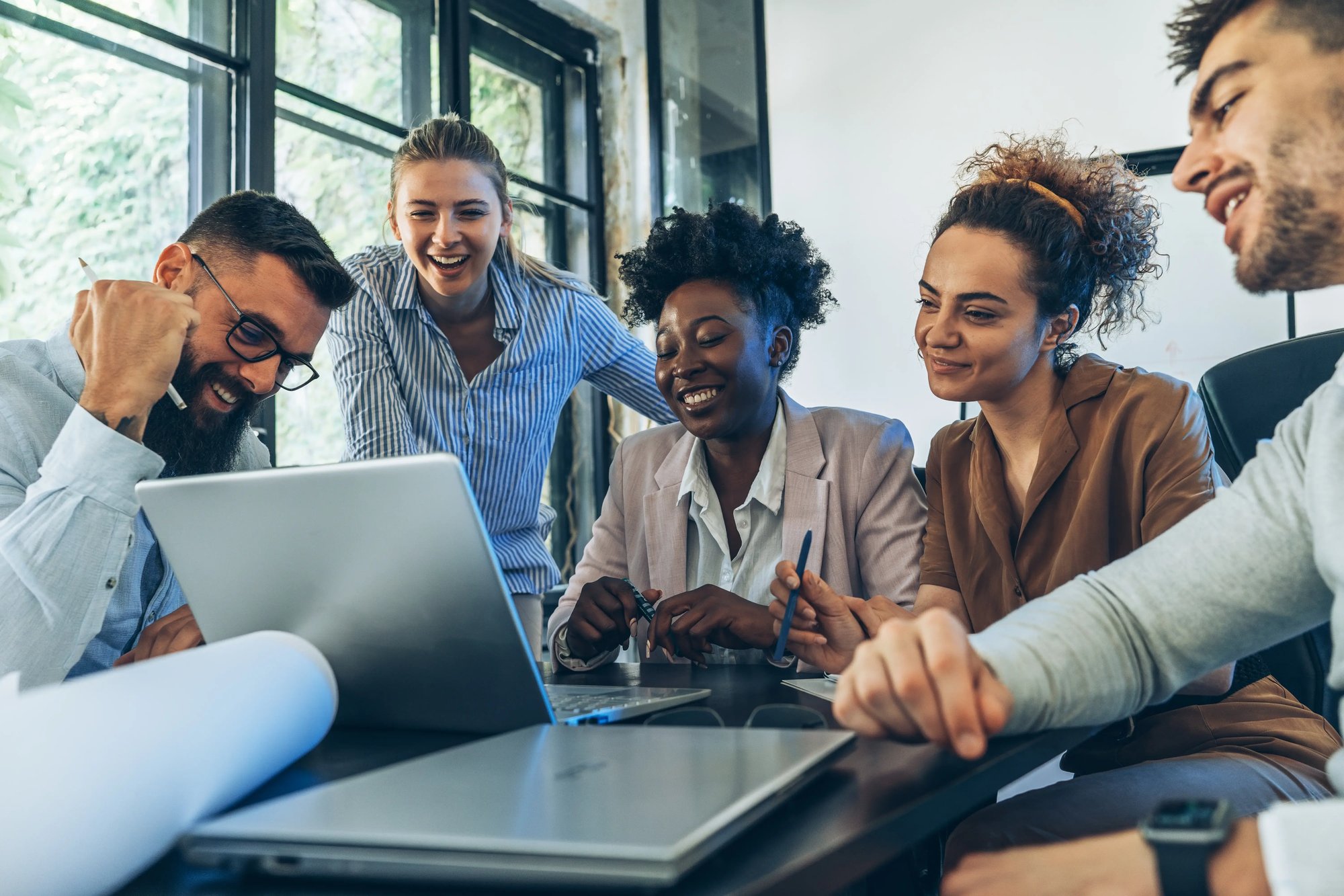 A diverse group of five young professionals gather around a laptop in a modern office, smiling and engaged in a collaborative discussion about discounted health assessments. Sunlight streams through large windows behind them.