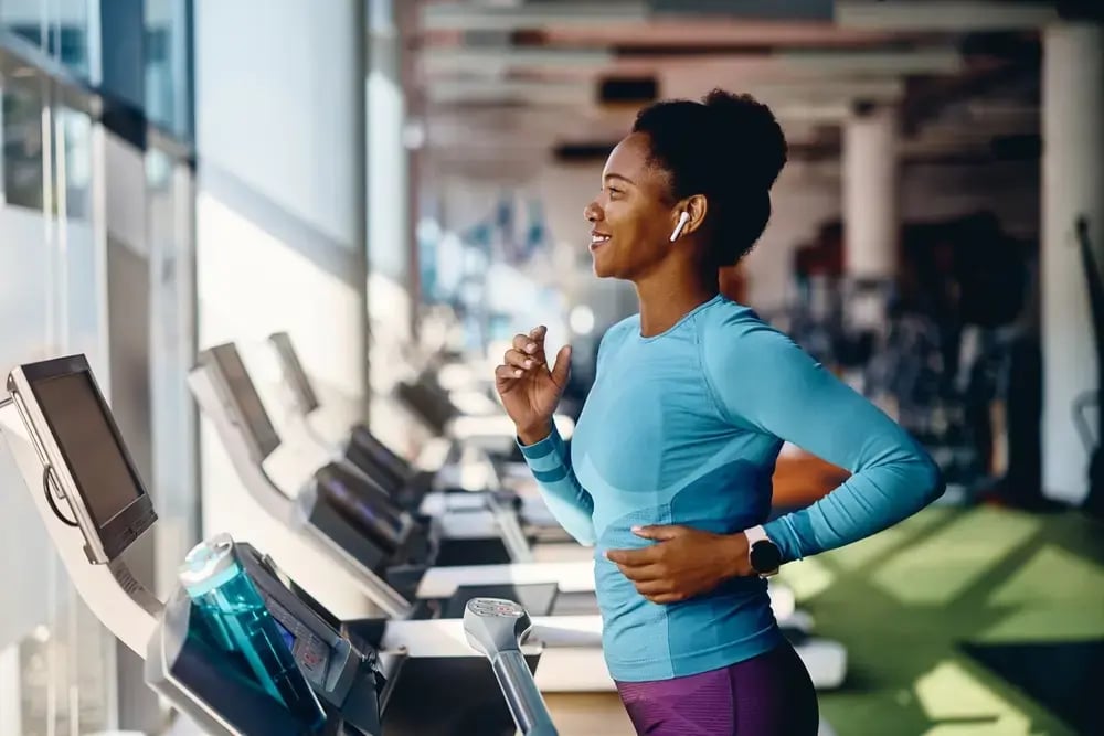 A woman in athletic wear smiles while running on a treadmill at a gym, wearing wireless earbuds and a smartwatch, as sunlight streams through large windows—embracing wellbeing at work through active, healthy habits.