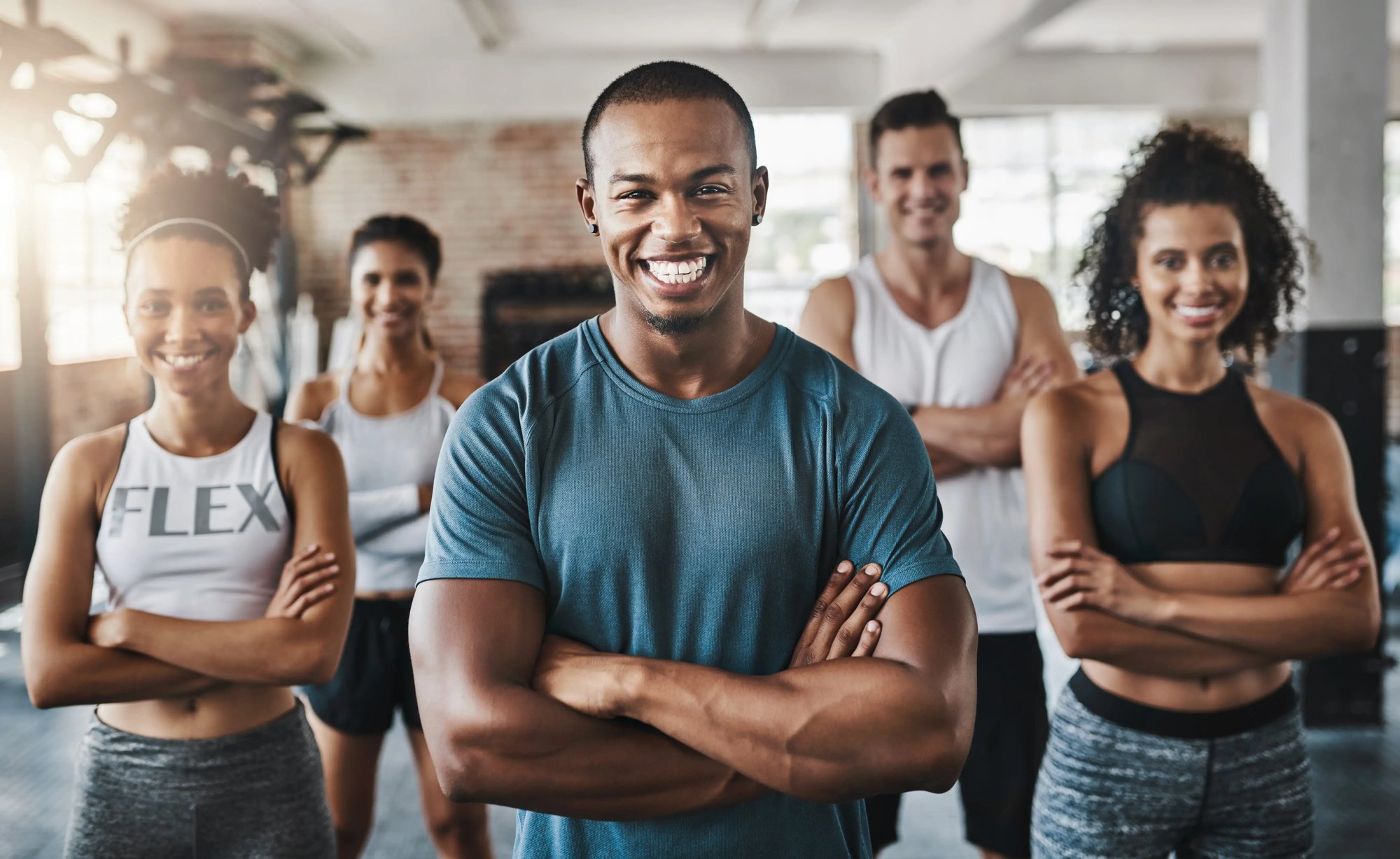 A group of people standing together in a gym, smiling and wearing athletic clothing. The scene showcases a diverse collection of individuals with varying body types and ages.