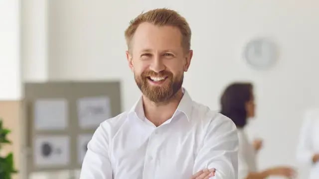 A man with a beard and light brown hair, wearing a white shirt, smiles with arms crossed in a bright office, reflecting feeling so much healthier thanks to his employee wellbeing benefits.