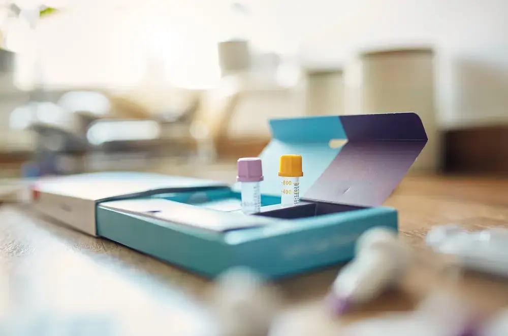 Two small vials with colored caps, resembling those found in at-home test kits, are placed inside an open blue box on a wooden surface, set against a blurred background of a home interior.