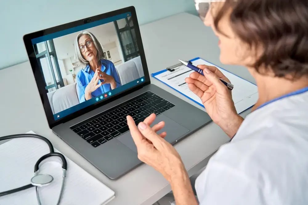 A healthcare professional sits at a desk with a stethoscope and clipboard, holding a pen while having a remote GP appointment with an older woman displayed on the laptop screen.