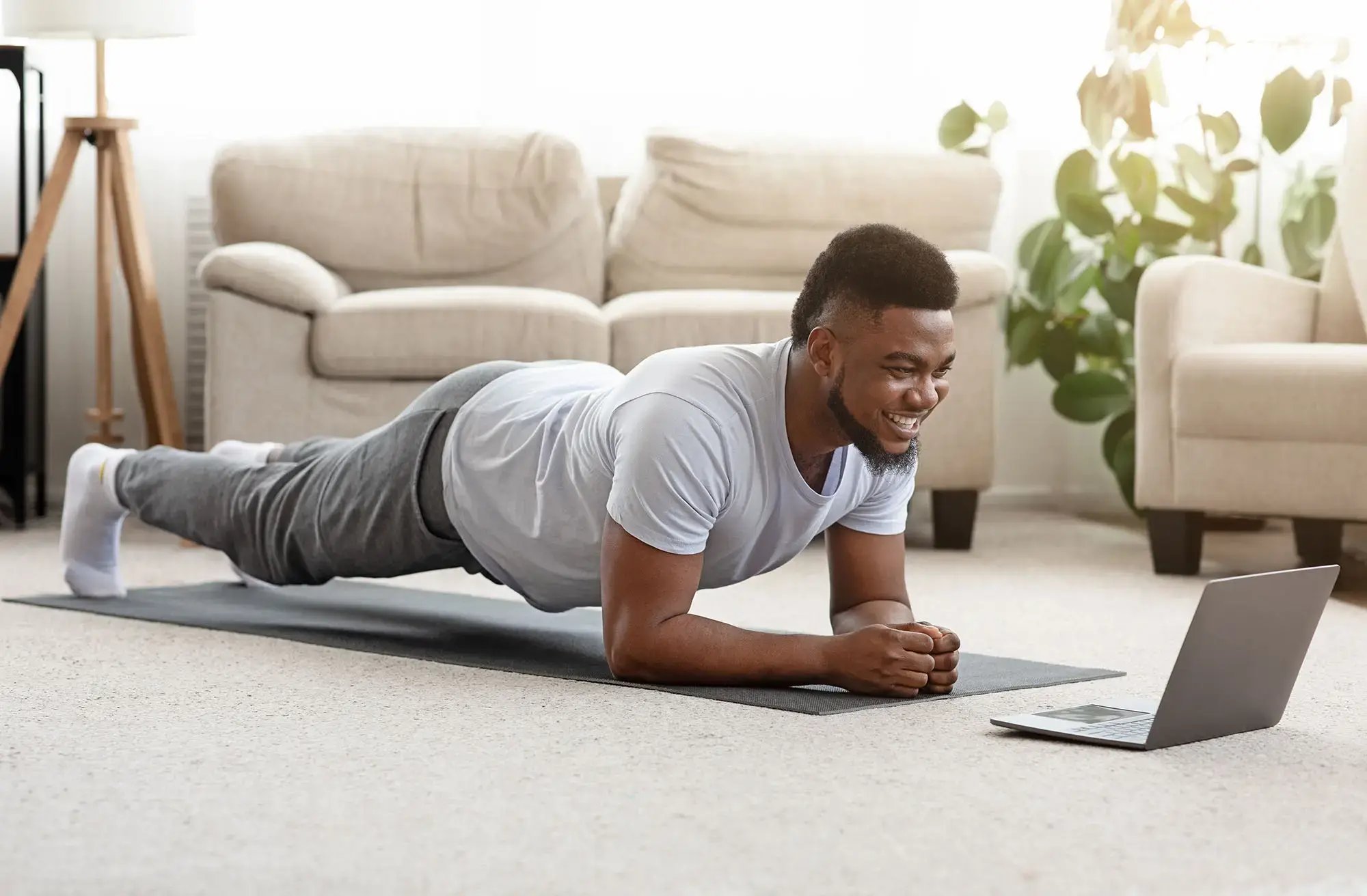 A man is performing a plank exercise on a mat in front of a laptop, surrounded by indoor furniture such as a couch. The setting features a wall and floor that contribute to the comfortable atmosphere.