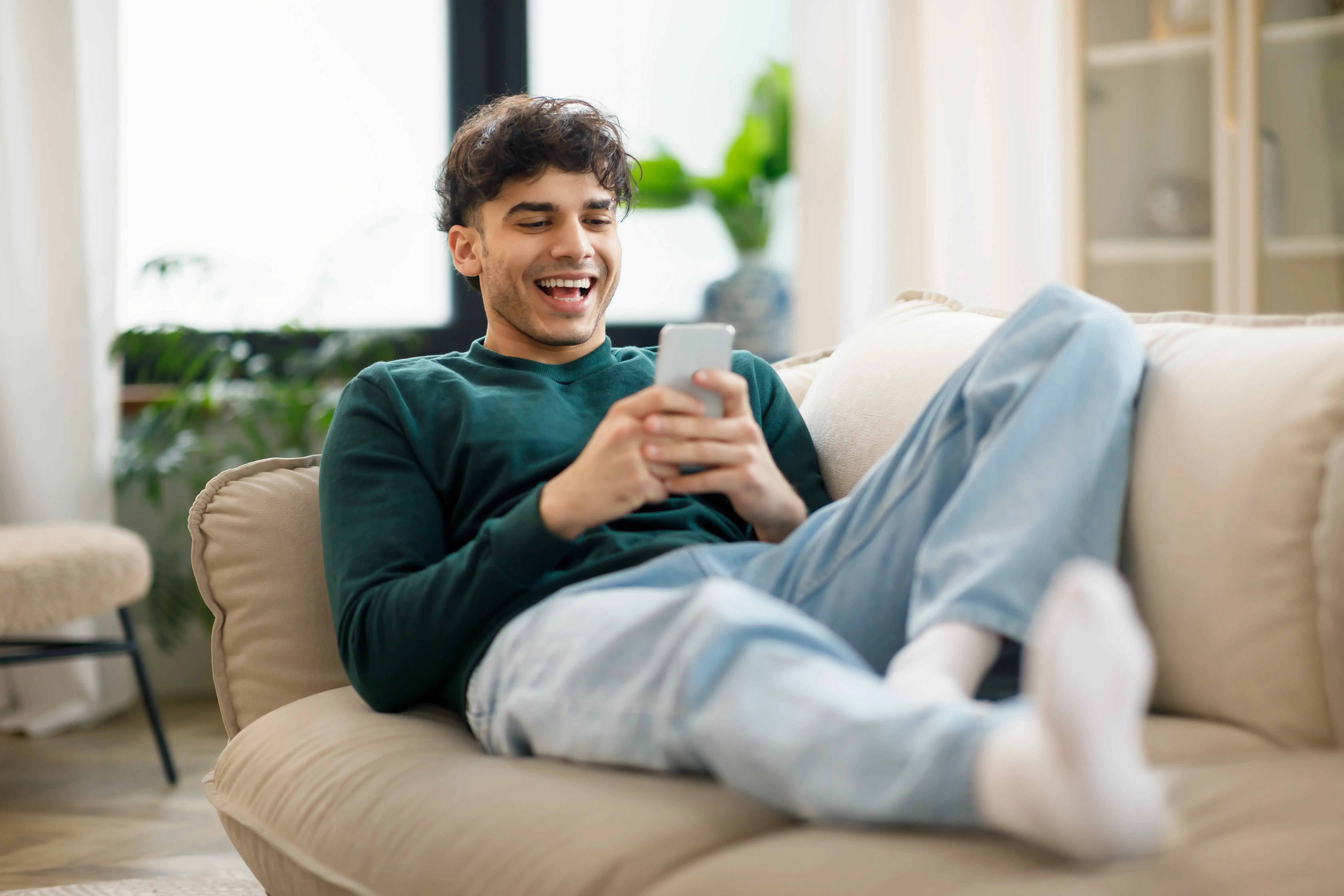 A man lying on a couch, looking at his phone with a smile, during an online counselling session in a comfortable indoor living room setting.