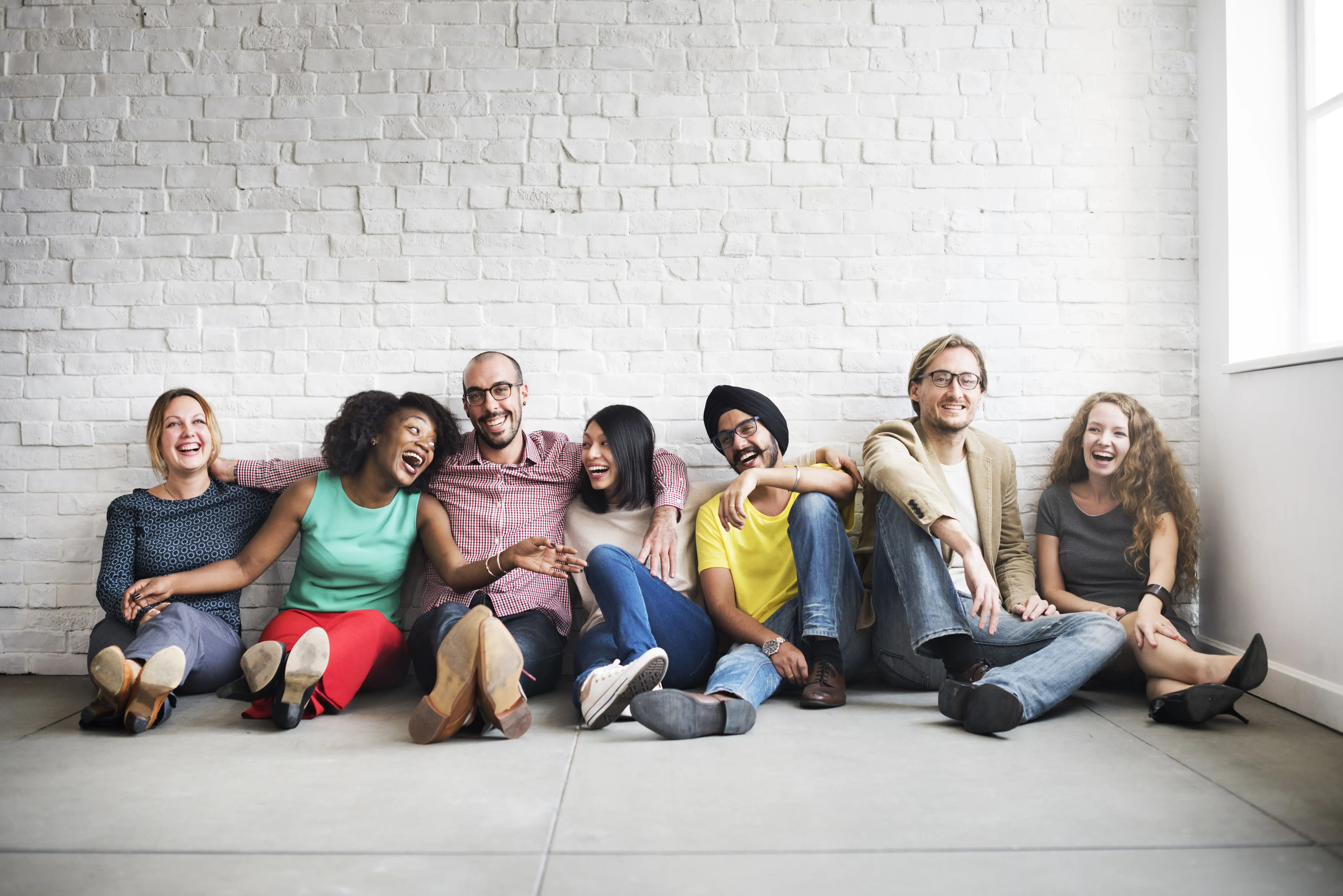 A diverse group of seven people sits on the floor against a white brick wall, smiling and laughing together. They appear relaxed and happy, enjoying each others company.